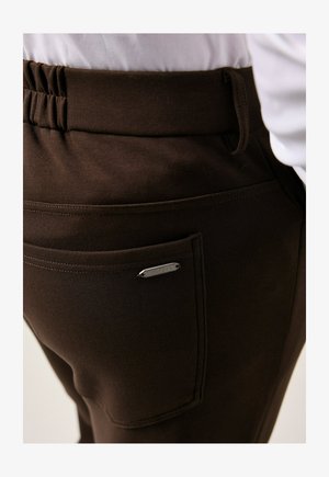 Close-up of dark brown pants with elastic waistband and back pocket featuring a small metal label with text "JP 1880".