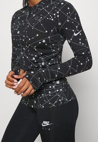 Schwarzes langärmliges Sportoberteil mit einem silbernen geometrischen Muster, Reißverschluss vorne und Daumenlöchern. Kombiniert mit schwarzen Leggings, die das "Nike Air"-Logo zeigen.
