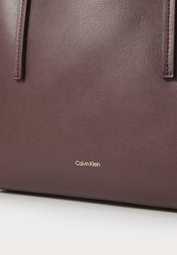 Sac à main en cuir bordeaux foncé avec deux anses plates, texture lisse, et un logo "Calvin Klein" doré embossé en bas.