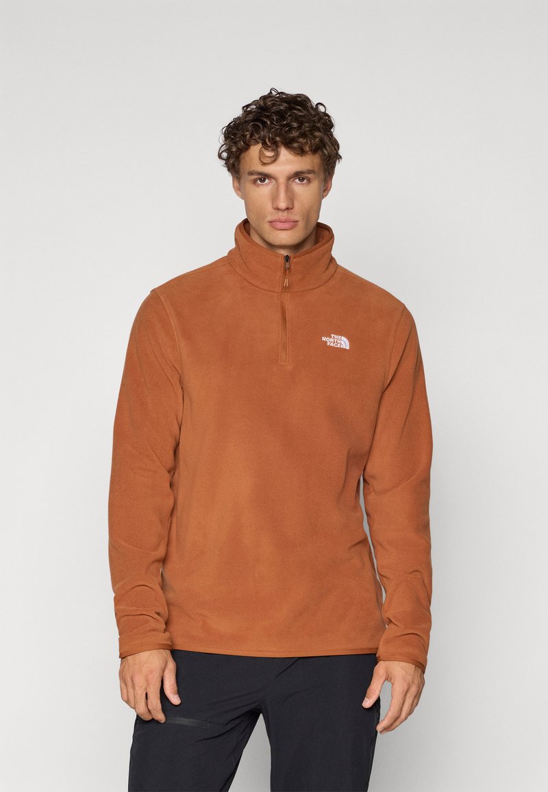 Pull en polaire couleur orange brûlé avec un col montant et une fermeture éclair mi-haute. Présente un logo blanc sur la poitrine gauche. Texture douce.