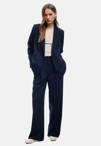 Marineblaues, doppelt geknöpftes Blazer mit vertikalen Nadelstreifen, kombiniert mit maßgeschneiderten Hosen und einem hellbeigen Oberteil. Mit subtilen Details akzentuiert.
