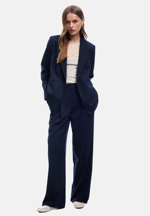 Marineblauwe dubbelgeknopte blazer met verticale streepjes, gecombineerd met op maat gemaakte broeken en een lichtbeige top. Verfraaid met subtiele details.