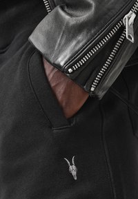 Sudadera negra con mangas de cuero, presenta un bolsillo lateral y un logotipo de calavera de cabra bordado en blanco en la parte frontal.