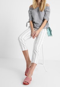 Off-shoulder randig blus i blått och vitt, parat med vita byxor med tunna vertikala linjer och rosa foppatofflor.