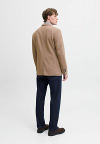 Beige gewebter Blazer mit Reverskragen und geknöpften Manschetten, kombiniert mit dunklen Jeans und braunen Schuhen, von hinten betrachtet.