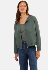 Zelená bomber bunda s elastickými manžetami, dvěma předními kapsami a zipovým zapínáním. Nositá přes šedý top a v kombinaci s modrými džíny.