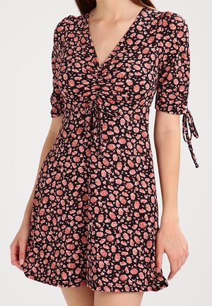 Robe noire avec motif floral rose, encolure en V froncée, manches courtes bouffantes avec liens et jupe évasée portée par un mannequin.