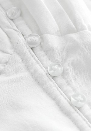 Bílý textil s hladkou strukturou, zdobený dvěma jasnými knoflíky podél švu. Tkanina je lehká a mírně pomačkaná.