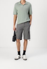 Man med ljusgrön kortärmad stickad topp, grå cargoshorts, svarta strumpor, vita sneakers, som håller en liten svart väska i höger hand.