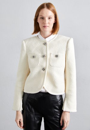 Chaqueta blanca texturizada con cuello redondo, dos bolsillos frontales, botones decorativos y mangas largas, combinada con pantalones de cuero negros.
