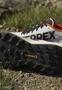 Chaussure de trail avec semelle en caoutchouc Continental noire, dessus blanc, lacets noirs et bordure orange reposant sur un sol rocheux.