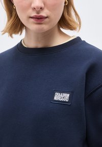 Γυναίκα που φοράει μπλε φούτερ με μικρό ορθογώνιο λευκό ετικέτα λογότυπου "NØRGAARD" στο στήθος, με ορατά χρυσά σκουλαρίκια κρίκους.