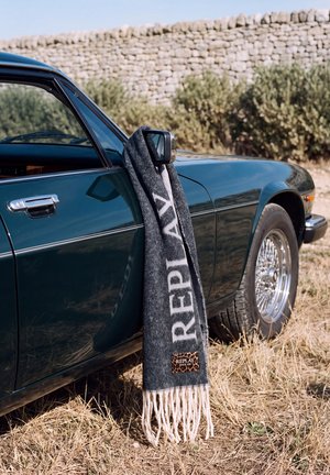 Une écharpe grise Replay est accrochée au rétroviseur latéral d’une voiture ancienne vert foncé garée près d’un mur de pierre.