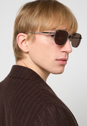 Giovane con capelli castano chiaro, che indossa occhiali da sole aviator con lenti scure e una giacca marrone scuro a righe sottili, che guarda di lato.