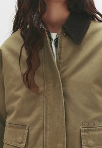 Veste en coton vert olive avec col en velours côtelé, fermeture éclair et deux poches avant, portée sur une chemise à carreaux noir et blanc.