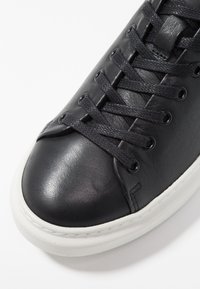 Baskets en cuir noir avec un bout arrondi, une tige texturée, des lacets plats noirs et une semelle en caoutchouc blanche. Elles disposent d'œillets métalliques et de coutures minimales.