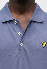 Sinist värvi teksuurse kangaga polo-särk, millel on krae, kaks nuppu ja rinnal tikitud kollane kotkalogo.