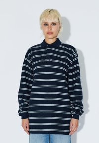 Navy striped long-sleeve polo shirt with white horizontal stripes, featuring a collar and relaxed fit, paired with light blue jeans.