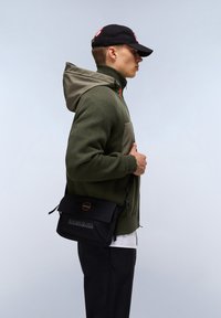 Felpa con cappuccio verde oliva e trama, abbinata a una borsa a tracolla nera con logo e chiusura a patta. Cappellino nero indossato.