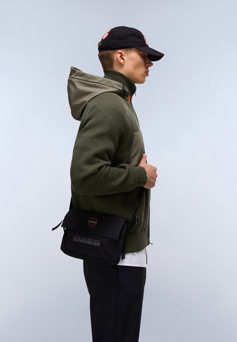 Felpa con cappuccio verde oliva e trama, abbinata a una borsa a tracolla nera con logo e chiusura a patta. Cappellino nero indossato.