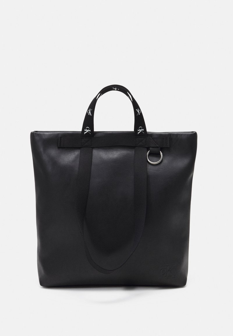 Sac fourre-tout en cuir noir avec deux anses en tissu imprimées "CK" et un détail en anneau métallique argenté sur un fond uni.