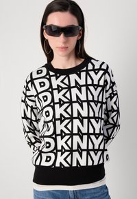 Juodas ir baltas megztinis su drąsiais geometriniais raštais ir "DKNY" logotipu; su raukinėta apykakle ir juosmeniu, minkšta tekstūra ir laisvas siluetas.