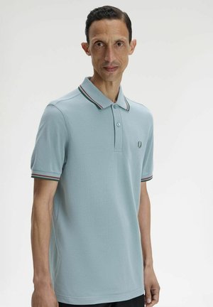 Hombre con camiseta polo de manga corta azul claro con cuello y puños de las mangas a rayas, posando frente a un fondo blanco liso.