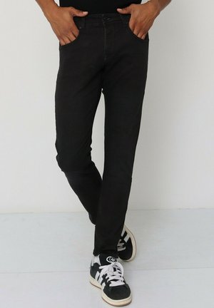 Jean slim noir avec cinq poches et une texture lisse, associés à des chaussures de sport noir et blanc avec des lacets et des rayures contrastées.