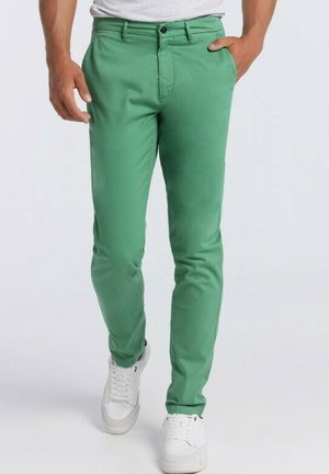 Chinos en coton vert avec une coupe ajustée, poches avant et fermeture par bouton. Associés à des baskets blanches. Texture lisse et couleur unie.
