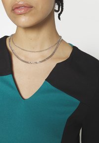 Personne portant un haut col en V bleu sarcelle et noir et deux colliers chaîne en argent, lèvres légèrement entrouvertes, cheveux tressés visibles.