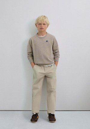 Pulover gri deschis cu manșete cu nervuri, pantaloni chinos bej și adidași maro cu accente galbene. Copilul stă drept în fața unui fundal simplu.