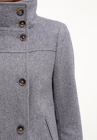 Abrigo de lana gris con un cuello alto, cuatro botones oscuros en el frente y dos bolsillos laterales. Presenta una textura suave y un diseño entallado.