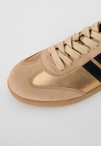 Beige atletická obuv s zlatým metalickým panelem, černým pruhem, texturovaným semišem a tkanými šňůrkami na lehké podrážce.