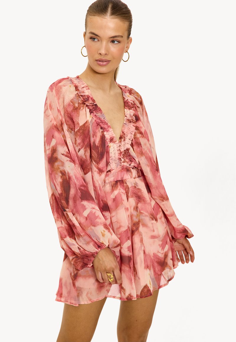 Vrouw in een roze en rode bloemenjurk met lange mouwen en een gerimpelde V-hals, met gouden ringoorbellen, poseert tegen een effen witte achtergrond.