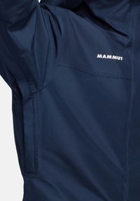 Mammut-Jacke in Marineblau. Sie verfügt über einen glatten, wasserdichten Stoff, Reißverschlusstaschen und ein markantes Logo im oberen Bereich.