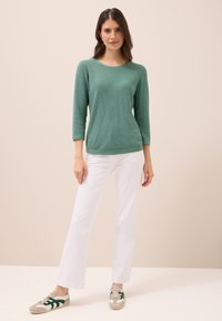 Grüner, strukturierten Strickpullover mit Dreiviertelärmeln, kombiniert mit weißen Schlaghosen und beigen Turnschuhen mit grünen Akzenten.