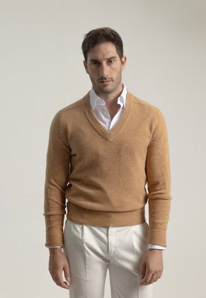 Suéter de lana marrón claro con escote en V y puños acanalados. Llevado sobre una camisa blanca con cuello, combinado con pantalones color crema.