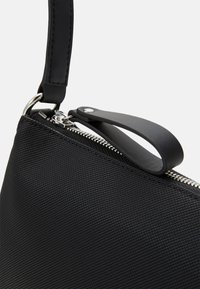 Bolsa de tela texturizada negra con una correa de cuero suave, herrajes de cremallera plateados y un lazo lateral que facilita el manejo.