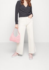 Sweter w ciemnoszarym kolorze, w stylu narzutki, białe szerokie spodnie dżinsowe oraz różowa pleciona torebka. Modelka nosi różowe sneakersy, pozując na jednolitym tle.