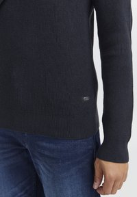 Marineblå strikket sweater med ribbet tekstur, tætsiddende manchetter og et lille mærke ved sømmen; parret med mørke denimjeans.
