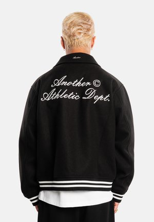 Persona con cabello rubio corto que lleva una chaqueta negra con puños a rayas blancas y el texto "Another Athletic Dept." bordado en la espalda.