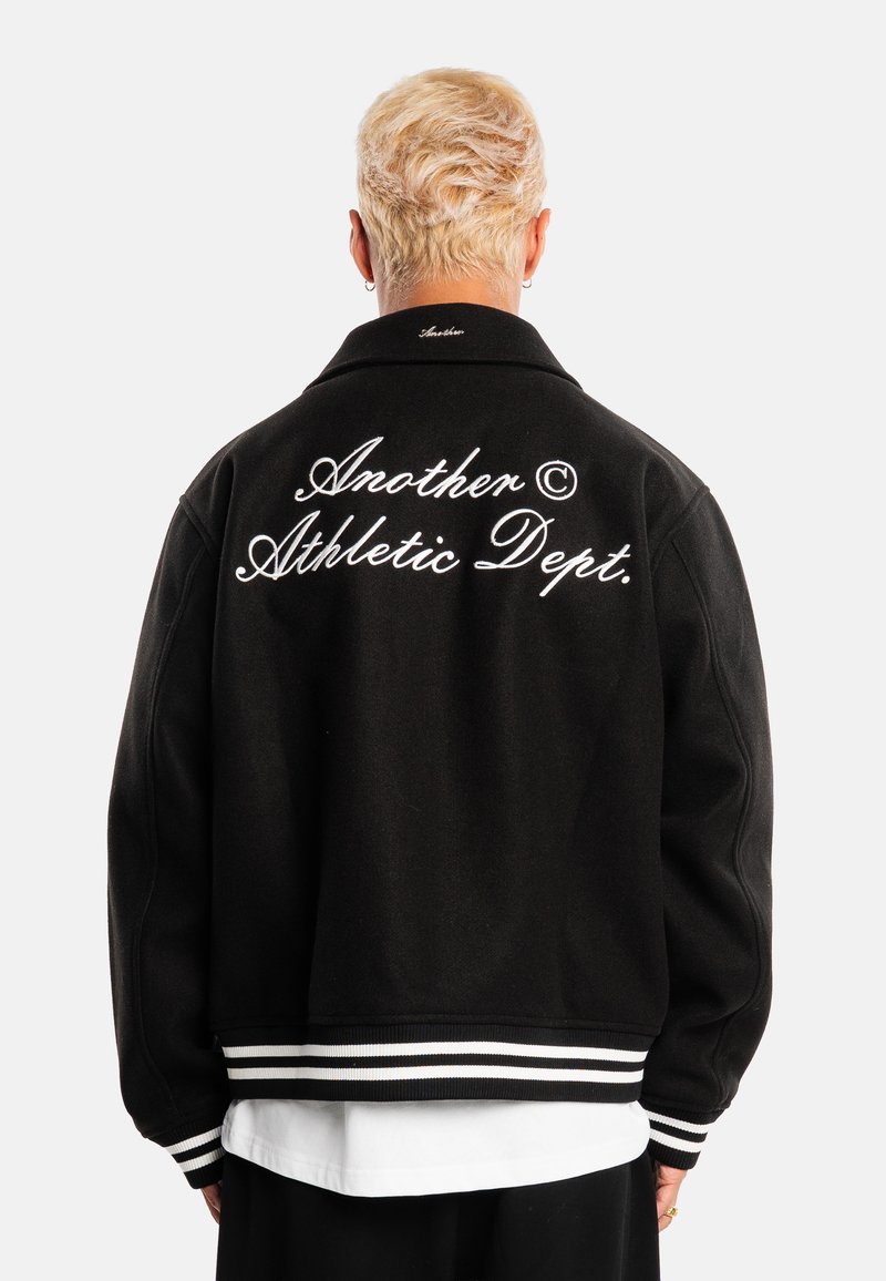 Personne aux cheveux blonds courts portant une veste noire avec des poignets rayés blancs et le texte "Another Athletic Dept." brodé dans le dos.