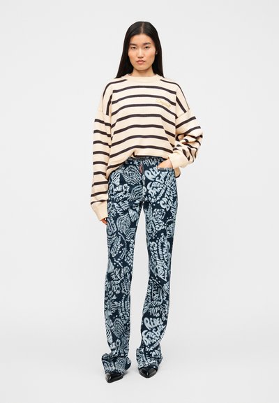 Pull beige à rayures avec une coupe oversize et des épaules tombantes, associé à un pantalon bleu orné d'un imprimé style graffiti. Des chaussures foncées complètent la tenue.
