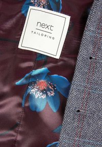 Witte "next TAILORING" label op bloemen maroon stof binnenin een grijze visgraatpatroon jacket met rode en blauwe stiksels.