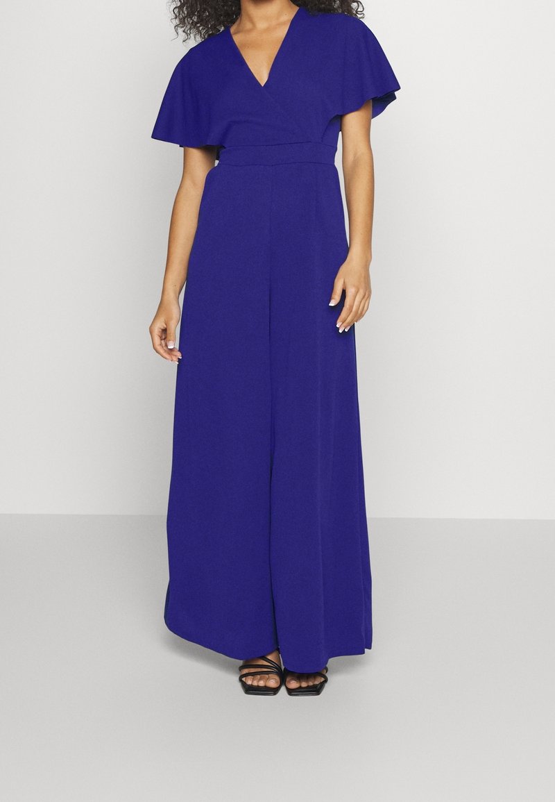 Mono azul con escote en V, mangas cortas y fluidas, y cintura ajustada. La tela es suave y se adapta a pantalones de pierna ancha. Tacones negros con tiras.