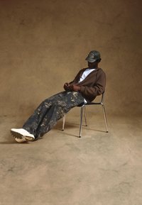 Brown oversized hoodie, white t-shirt, camouflage pants, green cap, seated on a chrome chair with white sneakers on a textured floor.