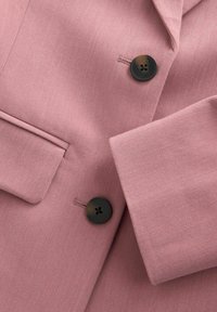 Roze blazer met een gladde stof, een reverskraag met inkeping en twee zwarte knopen aan de voorkant. Inclusief een zijzakdetail.
