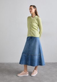 Zielony sweter z prążkowanym wzorem i guzikiem, niebieska dżinsowa spódnica w kształcie litery A oraz różowe teksturowane buty wsuwane. Swobodna, warstwowa stylizacja w kontrastowych kolorach.