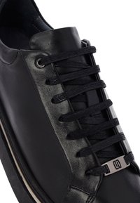 Sneaker in pelle nera con lacci neri infilati attraverso occhielli metallici e un'etichetta decorativa argentata, su uno sfondo bianco.
