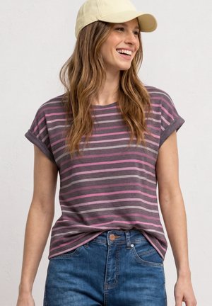 Femme souriante aux longs cheveux portant une casquette beige, une chemise rayée violette et rose aux manches retroussées et un jean bleu sur un fond uni.